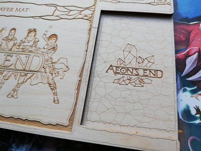 May include: A wooden game board with a light brown finish. The board has a raised section with the words "AEONS END" and a design of crystals. The board is part of a larger game set.