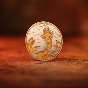 Puede incluir: Una moneda redonda de plata con un diseño dorado de un monstruo marino con la boca abierta. La moneda está sobre una superficie de madera marrón.