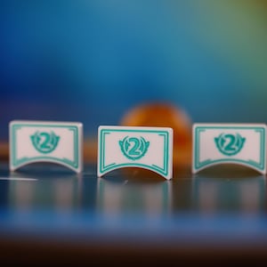 Könnte beinhalten: Drei weiße Spielmarken mit türkisfarbenen Akzenten und der Zahl "2" sind in einer Reihe angeordnet. Jede Marke hat einen türkisfarbenen Rand und ein stilisiertes Design. Der Hintergrund ist ein verschwommener Farbverlauf aus Blau, Orange und Gelb.