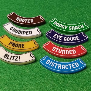 May include: Eight curved tokens with various words, including "ROOTED", "CHOMPED", and "PRONE", set against a green background. The tokens are in different colours, with white text. Other words include "DODGY SNACK" and "EYE GOUGE".