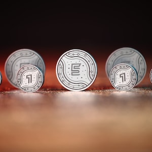 Könnte beinhalten: Sechs silberfarbene Token mit einem Leiterplatten-Design. Der mittlere Token hat den Buchstaben "E" in der Mitte. Die anderen fünf Token haben die Zahl "1" in der Mitte.