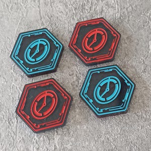 Op de afbeelding: Vier zwarte, zeshoekige tokens met een rood of blauw ontwerp. Elke token heeft een gekleurde omtrek en een centraal ontwerp dat lijkt op een klok of kompas. De tokens zijn in een vierkant patroon gerangschikt.