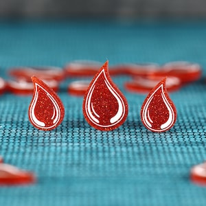 May include: Three red glitter blood drop shaped tokens with white outlines on a blue textured surface.