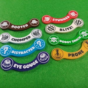 May include: A collection of colorful, curved game tokens with various status effects printed on them. The tokens are in shades of brown, red, white, blue, green, and yellow. Each token has a unique icon and text, such as "Rooted" or "Stunned".