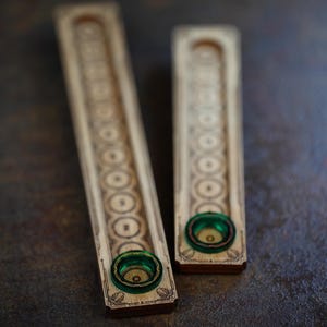 Può includere: Due porta incenso in legno con intricati disegni circolari e floreali. Ogni supporto ha un piccolo inserto rotondo di colore verde. I supporti sono rettangolari e di colore legno chiaro.