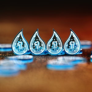 Op de afbeelding: Vier blauwe, traanvormige tokens met een wit ontwerp en het nummer "7" op elk. De tokens zijn in een rij op een bruin oppervlak gerangschikt.