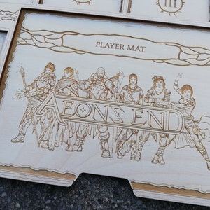 May include: A wooden player mat for the board game "Aeon's End". The mat features a light brown background with a decorative border and a laser-etched image of six characters standing in front of the game's title.