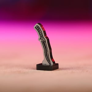 May include: A black and silver miniature dagger with a red blade, standing on a black base.