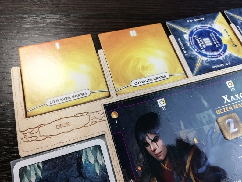 May include: A wooden game organizer with two yellow cards that say "OTWARTA BRAMA" and a character card with a woman with dark hair and a red and black outfit. The character card has the text "XAXC" and "UCZEŃ MAG" on it.