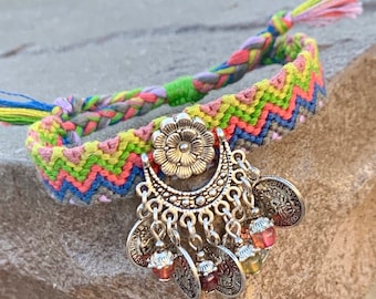 Pode incluir: Um bracelete de amizade colorido com um amuleto de prata com uma flor e moedas penduradas. O bracelete é feito com um padrão de chevron e tem um esquema de cores rosa, verde, amarelo e azul.