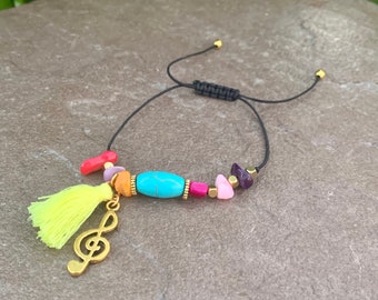 Pode incluir: Uma pulseira colorida com contas e um cordão preto ajustável. A pulseira apresenta uma conta turquesa, pedras rosa e roxas e um berloque de clave de sol dourada. Uma borla amarela adiciona um toque divertido ao design.