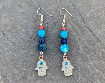 Pode incluir: Brincos de ouro com um design de contas de pedras preciosas azuis e vermelhas e um amuleto de mão de hamsa com um olho turco azul no centro.