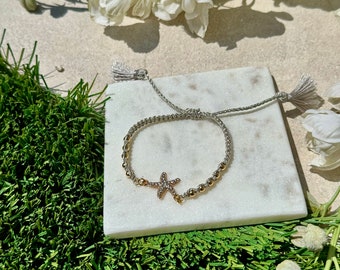 Pode incluir: Um bracelete de contas prateadas e douradas com um fecho de estrela do mar. O bracelete tem um fecho de nó deslizante e duas borlas.