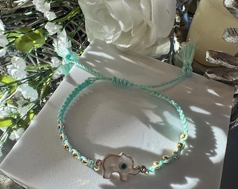 Pode incluir: Uma pulseira trançada azul clara com um berloque de elefante dourado, uma incrustação branca e um pequeno olho azul. A pulseira também tem contas douradas e pontas de borla. A pulseira é exibida em uma superfície branca.