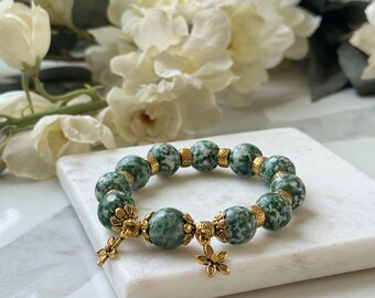 Pode incluir: Um bracelete de pedra de ágata verde e branca com detalhes dourados e pingentes em forma de flor. O bracelete está sobre uma superfície de mármore branco.