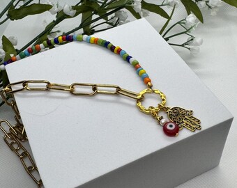 Pode incluir: Um colar de corrente dourada com uma fileira de contas coloridas e um pingente dourado com uma mão de Hamsa, um olho vermelho e uma pequena pérola branca. O colar é exibido em uma superfície branca com flores brancas ao fundo.