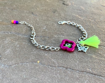 Pode incluir: Um bracelete de corrente prateada com um amuleto de flor rosa com um olho turco verde, um amuleto de borboleta prateada e um tassel verde.