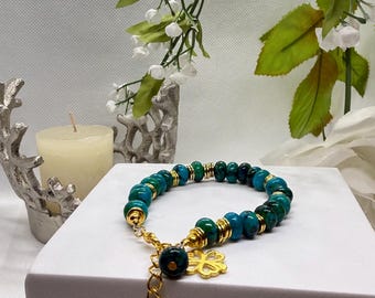 Pode incluir: Uma pulseira de contas turquesa e verdes com detalhes dourados. A pulseira tem uma corrente dourada e um berloque de trevo de quatro folhas. A joia é exibida em uma superfície branca com detalhes florais.