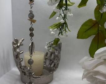 Pode incluir: Um castiçal prateado com uma vela branca e um enfeite de contas pendurado. O enfeite apresenta um pingente de pena e várias contas. Flores brancas e vegetação ao fundo.