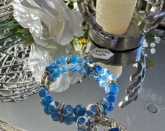 Pode incluir: Uma pulseira de contas azuis e brancas com detalhes dourados e um berloque. A pulseira é feita de contas redondas de ágata azul e branca. O berloque é prateado com uma pedra azul. A pulseira é exibida em uma superfície espelhada com uma vela e flores.