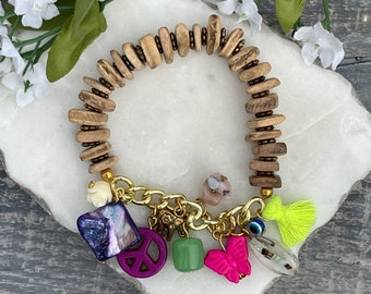 Pode incluir: Um bracelete de contas de madeira com uma corrente de ouro e vários pingentes, incluindo um sinal de paz, uma borboleta, um mau-olhado e uma borla. Os pingentes são de várias cores, incluindo roxo, rosa, verde e azul.