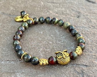 Pode incluir: Um bracelete de pedras preciosas verdes e vermelhas com detalhes dourados e um amuleto de coruja. O bracelete é feito com pequenas contas redondas e tem um amuleto de folha dourada de cada lado da coruja.
