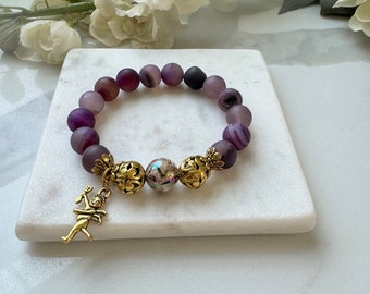 Pode incluir: Pulseira de contas de ágata roxa com detalhes dourados e um pequeno amuleto de querubim. A pulseira está sobre uma superfície branca.