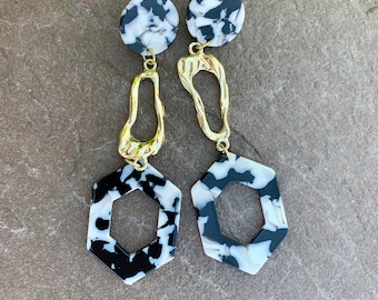 Pode incluir: Um par de brincos hexagonais com padrão de mármore preto e branco e detalhes dourados. Os brincos apresentam uma forma hexagonal com um recorte no centro e uma peça dourada pendente.