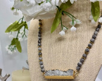 Pode incluir: Um colar com um pingente de pedra cinza emoldurado em uma moldura dourada. O colar apresenta contas cinza e douradas. O colar é exibido em um manequim bege. Flores brancas e vegetação ao fundo.