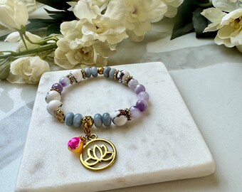 Pode incluir: Um bracelete feito de contas cinzas, brancas e roxas com um amuleto de lótus dourado. O bracelete está sobre uma superfície de mármore branco.