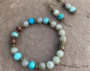 Pode incluir: Um bracelete de contas com um amuleto de Buda, contas marrons, azuis e cinzas, e brincos combinando com contas azuis e cinzas.