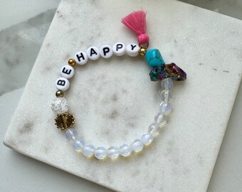 Pode incluir: Pulseira de contas brancas com as palavras "BE HAPPY" escritas em letras pretas. A pulseira tem um tassel rosa e um amuleto de cristal turquesa e iridescente.