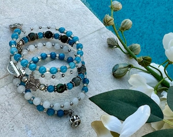 Pode incluir: Uma pulseira de várias voltas com contas azuis, brancas e verde escuro. A pulseira tem pingentes prateados, incluindo uma borboleta, uma folha e uma coroa. A pulseira é exibida em uma superfície cinza claro com um fundo de água azul.