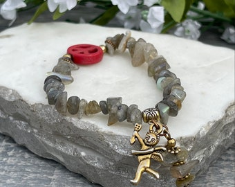 Pode incluir: Uma pulseira de pedras preciosas com pedras irregulares em tons de cinza. Um sinal de paz vermelho e um pingente dourado de uma figura alada são incorporados. A pulseira é exibida em uma superfície branca texturizada.