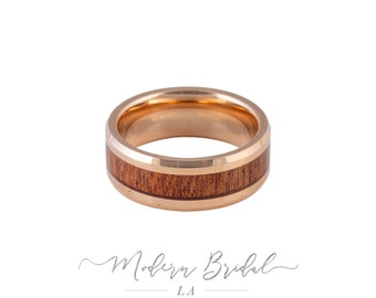 May include: A rose gold tungsten carbide ring with a brown wood inlay. The ring has a beveled edge and a polished finish.