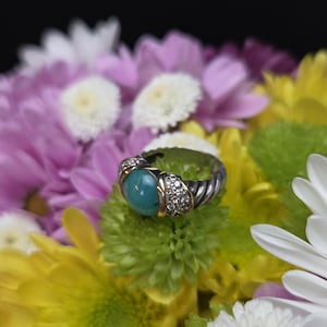 May include: A silver ring with a turquoise stone and diamond accents, set against a backdrop of colorful flowers. The ring features a twisted band design and gold-colored accents around the turquoise stone and diamond clusters.