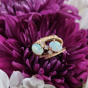 Könnte beinhalten: Ein goldener Ring mit zwei ovalen Opalen und kleinen Diamanten. Der Ring ist vor einem Hintergrund aus lila und weißen Blumen platziert. Das Design des Rings ist elegant und auffällig.