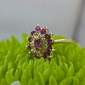 May include: A gold ring featuring a central oval-cut red gemstone surrounded by a cluster of smaller round-cut red gemstones and tiny clear accent stones. The setting is arranged in a floral-like pattern with a polished gold band.