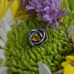 May include: A silver-toned metal bead featuring a swirling, knotted design with a single, round, faceted yellow gemstone set in the center. The bead is nestled within the petals of a bright green chrysanthemum flower with purple accents.