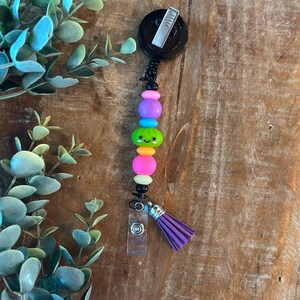 May include: A badge reel featuring a black circular housing with a silver metal clip. A vertical strand of colourful beads includes a green dumpling-shaped bead with a smiling face, flanked by pink, orange, blue, and black beads, ending in a purple tassel.