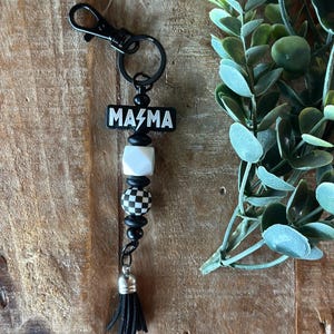 Può includere: Un portachiavi nero con una perlina rettangolare recante la scritta bianca "MA⚡MA", una perlina cubica bianca, una perlina rotonda a scacchi bianchi e neri e perline distanziatrici nere. Il portachiavi termina con una nappa nera e una chiusura in metallo nero.