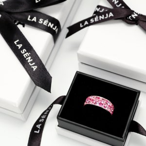 May include: A pink ring with small sparkling stones in a black ring box with a white lid. The box has a black ribbon with the text "LA S&Eacute;NJA" printed on it.