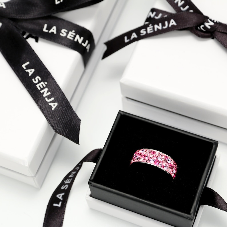May include: A pink ring with small sparkling stones in a black ring box with a white lid. The box has a black ribbon with the text "LA S&Eacute;NJA" printed on it.