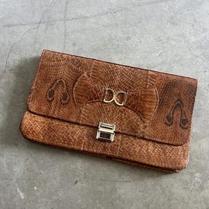 May include: Brown snakeskin-patterned clutch bag. The rectangular bag features a flap closure with a silver clasp and a gold-coloured "DC" emblem. The bag is made of a textured material and has a stitched design.