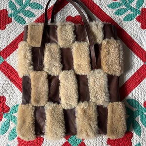 Peut inclure: Un sac fourre-tout marron et beige avec un motif en damier. Le sac présente une combinaison de cuir marron lisse et de patchs rectangulaires de matière beige et duveteuse. Il a deux poignées en cuir marron.