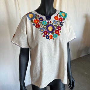 May include: Off-white, short-sleeved blouse with vibrant floral embroidery around the neckline. The embroidery features a variety of colorful flowers, including blue, orange, and purple. The blouse has a relaxed fit and a simple, elegant design.