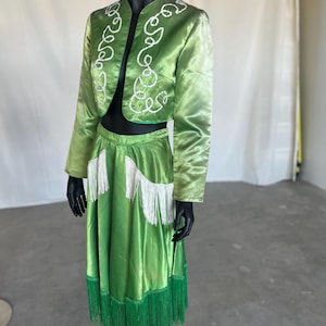 May include: A lime green two-piece outfit on a black mannequin. The jacket has white embroidered swirls and the skirt has white and green fringe. The outfit is made of a shiny, satin-like material.
