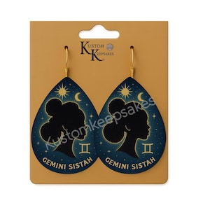 May include: Teardrop-shaped earrings with a dark blue background featuring a silhouette of a woman's profile, stars, and a crescent moon. The text "Gemini Sistah" is printed on the bottom. Gold-colored fish hook ear wires.