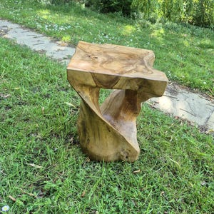 May include: A handcrafted wooden stool with a unique, organic design. The stool is made from a light brown wood with visible grain patterns. Its twisted base supports a square seat, creating a sculptural and functional piece. The stool is suitable for indoor or outdoor use.
