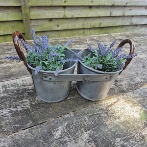 May include: A double metal bucket planter with two round containers connected by a central metal bar. Each bucket features a curved brown handle and contains artificial lavender sprigs with purple flowers and green stems in a grey finish.
